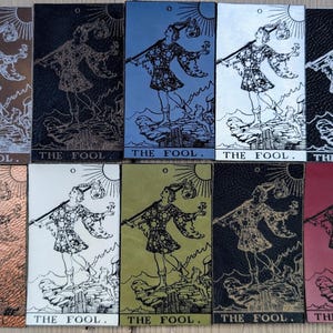 May include: A selection of tarot cards, each depicting "The Fool" in different colours. The cards show a detailed illustration of a figure with a staff, sun, and the text "THE FOOL." The cards have a textured finish and are arranged in a grid.