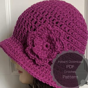 May include: A magenta crochet hat with a small brim and a large flower accent. The hat is handmade with a textured stitch pattern. The image includes text that says "Instant Download PDF Crochet Pattern".