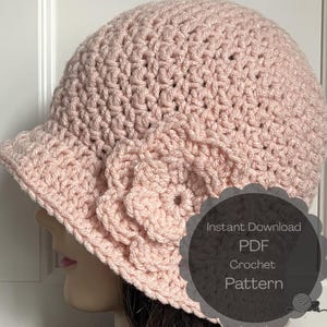 Puede incluir: Un sombrero cloche de ganchillo rosa claro con un adorno floral. El sombrero tiene un ala pequeña y un patrón de puntada texturizada. La imagen incluye un gráfico que dice "Instant Download PDF Crochet Pattern".