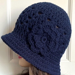 May include: A navy blue crocheted cloche hat with a floral detail. The hat has a small brim and an openwork pattern. The flower is on the side of the hat, adding a decorative touch.
