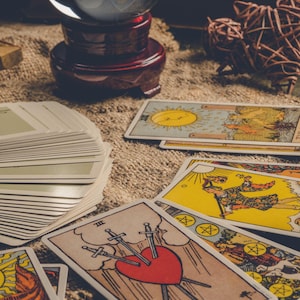 May include: A collection of tarot cards with various illustrations, including a sun, moon, and heart pierced by swords. A crystal ball and decorative objects are also present, suggesting a mystical theme.
