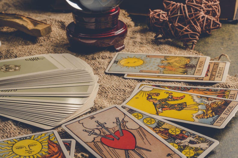 May include: A collection of tarot cards with various illustrations, including a sun, moon, and heart pierced by swords. A crystal ball and decorative objects are also present, suggesting a mystical theme.