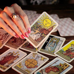 May include: A collection of tarot cards, featuring colorful illustrations and symbolic imagery, are arranged on a wooden surface. One card, depicting a sun, is held by a hand with a ring. Other cards show various figures and symbols.