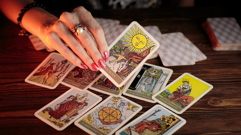 May include: A collection of tarot cards, featuring colorful illustrations and symbolic imagery, are arranged on a wooden surface. One card, depicting a sun, is held by a hand with a ring. Other cards show various figures and symbols.