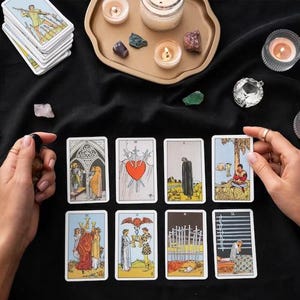 May include: A collection of tarot cards with colorful illustrations, including images of a heart pierced by swords and figures in robes. The cards are arranged on a black surface, alongside candles, crystals, and a decorative tray.