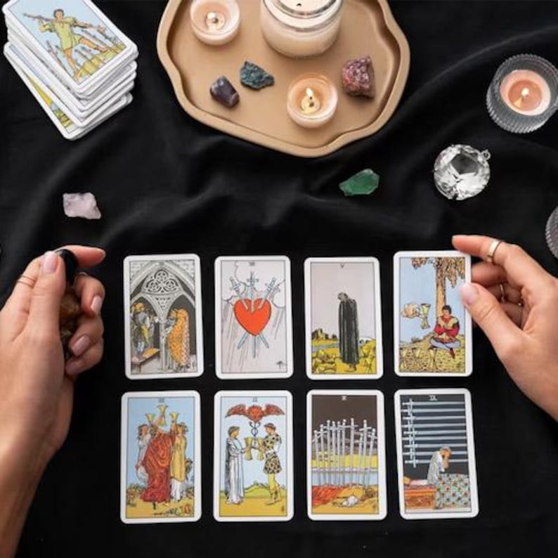 May include: A collection of tarot cards with colorful illustrations, including images of a heart pierced by swords and figures in robes. The cards are arranged on a black surface, alongside candles, crystals, and a decorative tray.