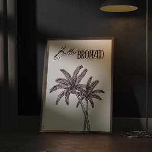 May include: A framed print featuring two palm trees in a black and white illustration. The text "Better BRONZED" is above the palm trees. The artwork is displayed on a dark surface, with a lamp and a rug in the background.