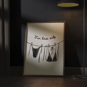 May include: A framed art print with a minimalist illustration of two bikinis hanging on a washing line. The phrase "Tan lines only" is written above the bikinis in a cursive font. The artwork is displayed in a dark room.