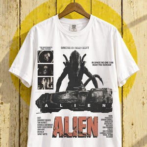 Peut inclure: Un t-shirt blanc avec un graphisme noir représentant la créature Alien et le vaisseau Nostromo. Le texte inclut 'DIRECTED BY RIDLEY SCOTT', 'IN SPACE NO ONE CAN HEAR YOU SCREAM', 'THE NOSTROMO'S FINAL VOYAGE', 'ALIEN', ainsi qu'une liste des acteurs et membres de l'équipe.