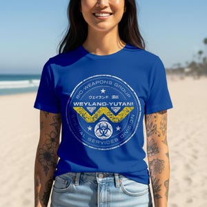 Puede incluir: Camiseta azul real con un diseño circular. El diseño incluye el texto "BIO-WEAPONS GROUP", "WEYLAND-YUTANI SPECIAL SERVICES DIVISION", un logotipo "W" amarillo y texto japonés. La camiseta es de cuello redondo y manga corta.