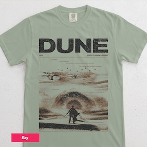 May include: Sage green t-shirt featuring the word "DUNE" in large, black block letters above a graphic from the novel by Frank Herbert. The graphic depicts a desert scene with figures and aircraft. The shirt also includes the text "1965" and "NOVEL BY FRANK HERBERT".