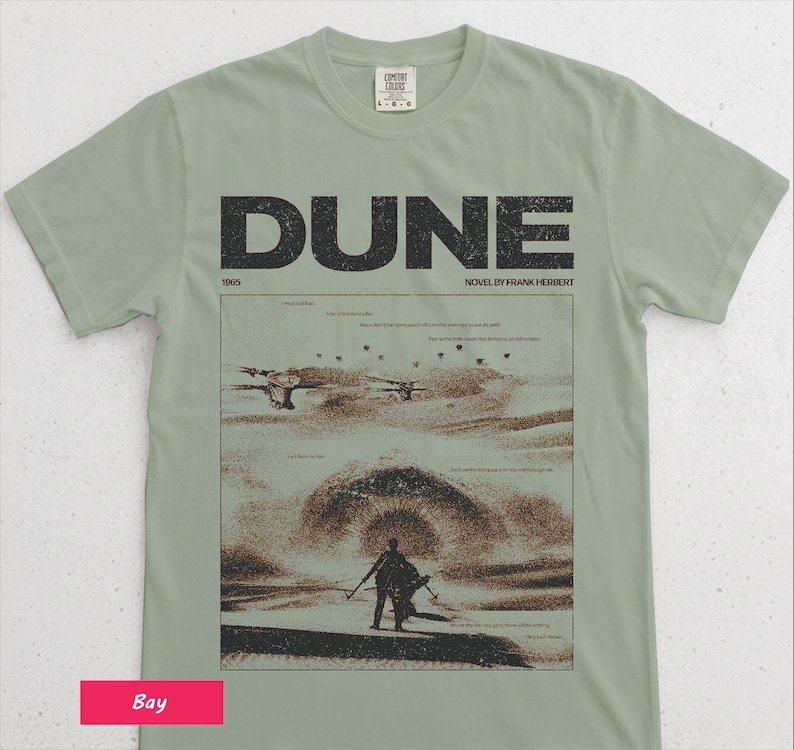 May include: Sage green t-shirt featuring the word "DUNE" in large, black block letters above a graphic from the novel by Frank Herbert. The graphic depicts a desert scene with figures and aircraft. The shirt also includes the text "1965" and "NOVEL BY FRANK HERBERT".