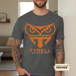 May include: Grey t-shirt featuring an orange owl-inspired design and the word "TYRELL". The text "MORE HUMAN THAN HUMAN!" is printed below. The shirt is a Comfort Colors Pepper tee.