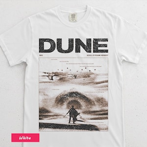 May include: White t-shirt featuring the word "DUNE" in large black letters, with the text "NOVEL BY FRANK HERBERT" below. The shirt displays a sepia-toned graphic from the novel, including a figure in a desert landscape. The year "1965" is also printed.