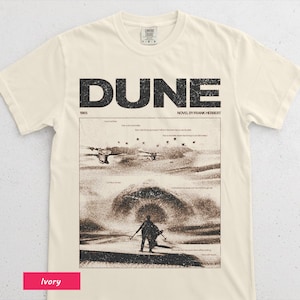 May include: Ivory-colored t-shirt featuring the word "DUNE" in large, black, block letters above a sepia-toned graphic from the novel by Frank Herbert. The graphic depicts a desert scene with figures and aircraft. The shirt is made of a soft material.