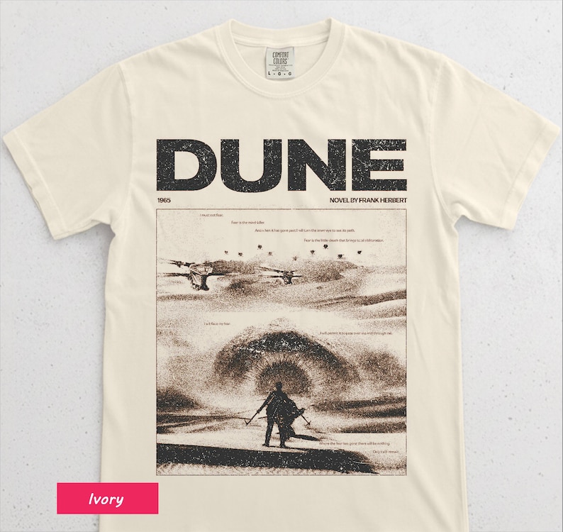 May include: Ivory-colored t-shirt featuring the word "DUNE" in large, black, block letters above a sepia-toned graphic from the novel by Frank Herbert. The graphic depicts a desert scene with figures and aircraft. The shirt is made of a soft material.