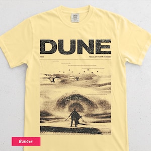 May include: Butter-colored t-shirt featuring the word "DUNE" in large, black letters above a sepia-toned graphic from the novel. The graphic depicts a desert scene with figures and text. The shirt also has the text "NOVEL BY FRANK HERBERT".