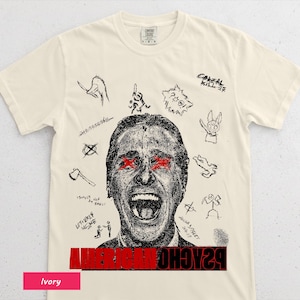 Könnte beinhalten: Elfenbeinfarbenes T-Shirt mit einer schwarz-roten Grafik eines schreienden Gesichts mit roten Augen, umgeben von Kritzeleien. Der Text "AMERICAN PSYCHO" ist unten rot aufgedruckt. Das Shirt ist aus weichem Material.