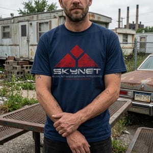 Puede incluir: Camiseta azul marino con un diseño geométrico rojo y la palabra "SKYNET" en letras blancas desgastadas. Debajo está el texto "NEURAL NET-BASED ARTIFICIAL INTELLIGENCE". Cuello redondo.