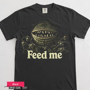 Puede incluir: Camiseta negra con una ilustración detallada de una planta carnívora y el texto "Feed me" en blanco roto. El diseño presenta una boca grande y dentada y detalles intrincados de la planta, creando un efecto visual llamativo.