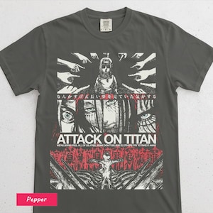 May include: A pepper-coloured t-shirt with a graphic from "Attack on Titan." The design features illustrations of characters, hands, and Japanese text. The text "ATTACK ON TITAN" is in white, with additional red text below.