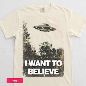 May include: Cream-coloured t-shirt with a retro design of a flying saucer above a forest. The words "I WANT TO BELIEVE" are printed in white below the image. The shirt is made of a soft material.