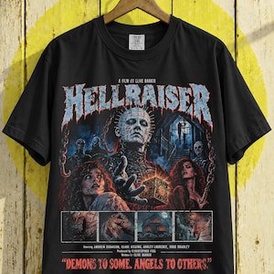May include: A black short-sleeved t-shirt featuring a graphic print from the film Hellraiser. The design includes Pinhead, the puzzle box, and other characters, with the text A FILM BY CLIVE BARKER and DEMONS TO SOME. ANGELS TO OTHERS. printed on the front.
