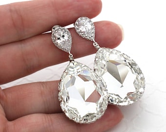 May include: Elegant, clear teardrop earrings with a sparkling, faceted design. The earrings have a smaller, round, crystal-studded top and a larger, pear-shaped crystal pendant. The earrings are held in a hand against a white background.