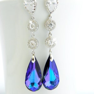 May include: Elegant silver-toned dangle earrings. Each earring features a pear-shaped clear crystal at the top, followed by three round, clear crystal accents, and a large, teardrop-shaped, iridescent blue-purple crystal at the bottom. The earrings are displayed against a white background.
