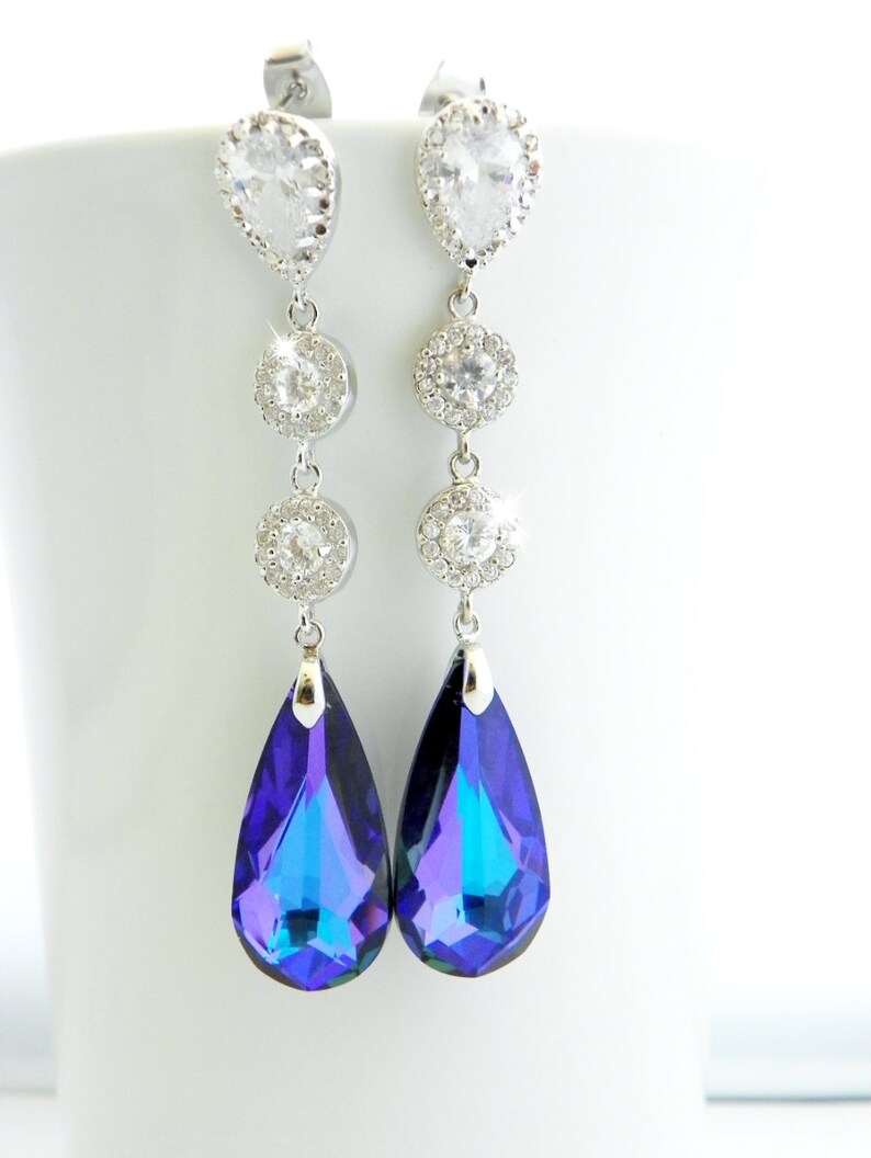 May include: Elegant silver-toned dangle earrings. Each earring features a pear-shaped clear crystal at the top, followed by three round, clear crystal accents, and a large, teardrop-shaped, iridescent blue-purple crystal at the bottom. The earrings are displayed against a white background.