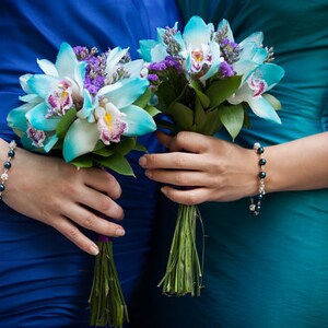 May include: Two bouquets of turquoise and white orchids with purple accents are held by two people wearing blue and teal dresses. Each person wears a bracelet with dark beads and silver accents. The bouquets have green stems.