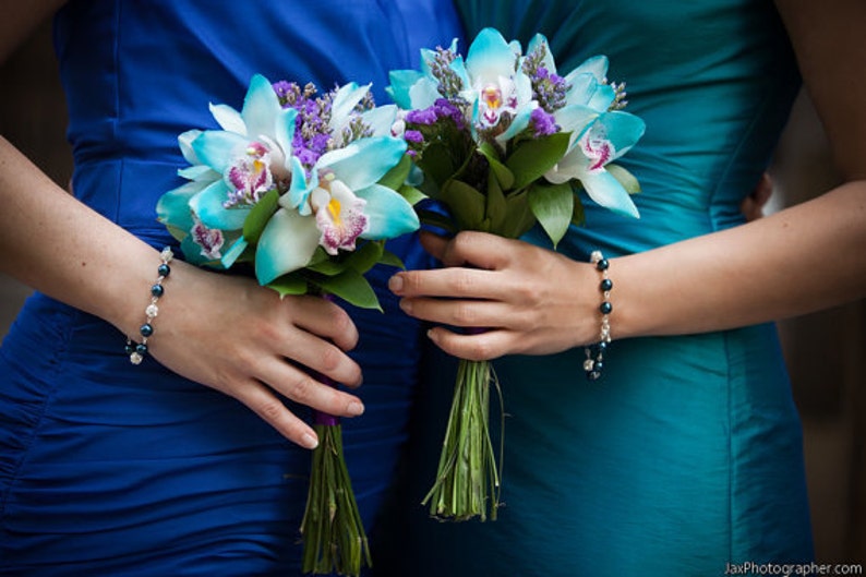 May include: Two bouquets of turquoise and white orchids with purple accents are held by two people wearing blue and teal dresses. Each person wears a bracelet with dark beads and silver accents. The bouquets have green stems.