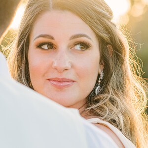May include: A woman with long, wavy blonde hair and brown eyes looks to the side. She wears earrings with a pearl and silver accents. The background is blurred with a warm, golden light.
