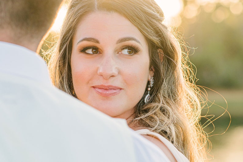 May include: A woman with long, wavy blonde hair and brown eyes looks to the side. She wears earrings with a pearl and silver accents. The background is blurred with a warm, golden light.