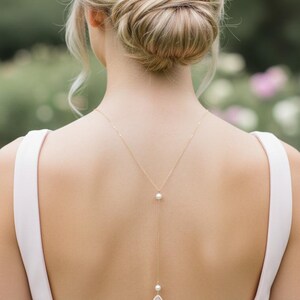 May include: A gold-colored necklace with a pearl and a teardrop-shaped pendant with a pink stone. The necklace hangs down the back of a person wearing a light pink dress. The person's hair is styled in a bun.