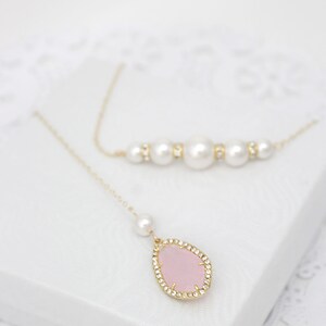 May include: Two necklaces on a white surface. One necklace features a series of large white pearls with gold accents. The other necklace has a gold chain with a pink teardrop pendant surrounded by small crystals and a pearl.