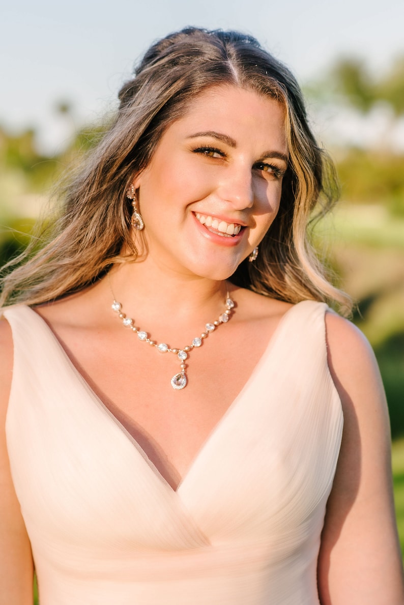 May include: A smiling person wearing a light pink dress and a sparkling necklace and earrings set. The necklace features a series of clear, round stones and a teardrop pendant. The earrings are a matching design. The background is blurred with green and yellow tones.