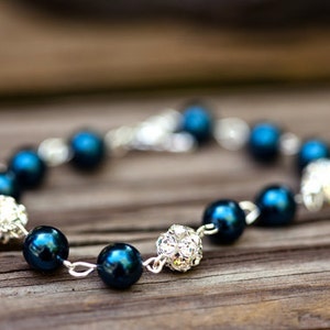 May include: A bracelet featuring dark teal beads and sparkling silver accents. The bracelet is strung on a silver chain and has a clasp closure. The beads are interspersed with clusters of small, glittering crystals.