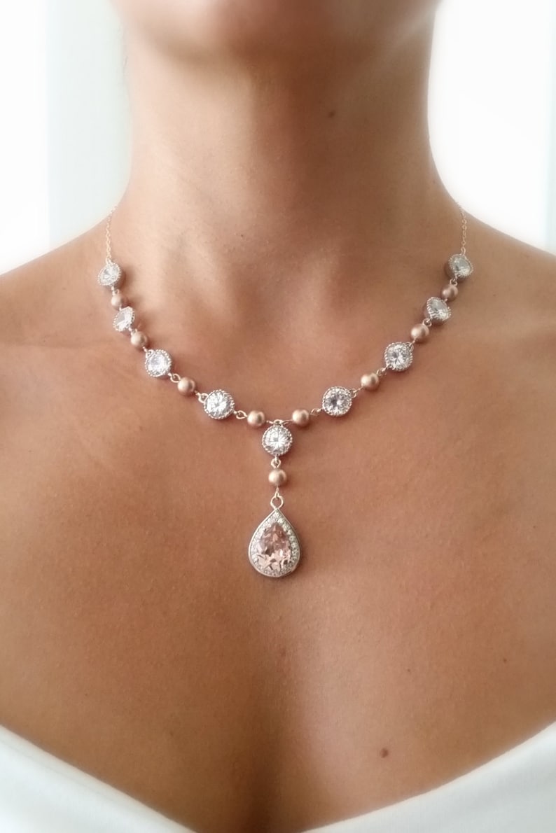 May include: A delicate necklace featuring a series of round, clear crystals set in a gold-toned metal. The necklace has a teardrop-shaped pendant with a large, light pink stone. The necklace is designed to be worn around the neck.