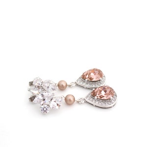 May include: Elegant silver-toned drop earrings. One earring features a cluster of clear crystals, while the other has a pear-shaped peach-colored gemstone surrounded by smaller clear crystals. A small, round, peach-colored bead hangs below the cluster and gemstone.