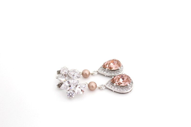 May include: Elegant silver-toned drop earrings. One earring features a cluster of clear crystals, while the other has a pear-shaped peach-colored gemstone surrounded by smaller clear crystals. A small, round, peach-colored bead hangs below the cluster and gemstone.