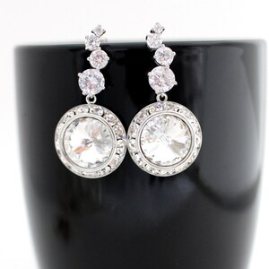 May include: A pair of silver dangle earrings with a large round clear crystal stone at the bottom. The earrings have a row of smaller clear crystal stones above the large stone.