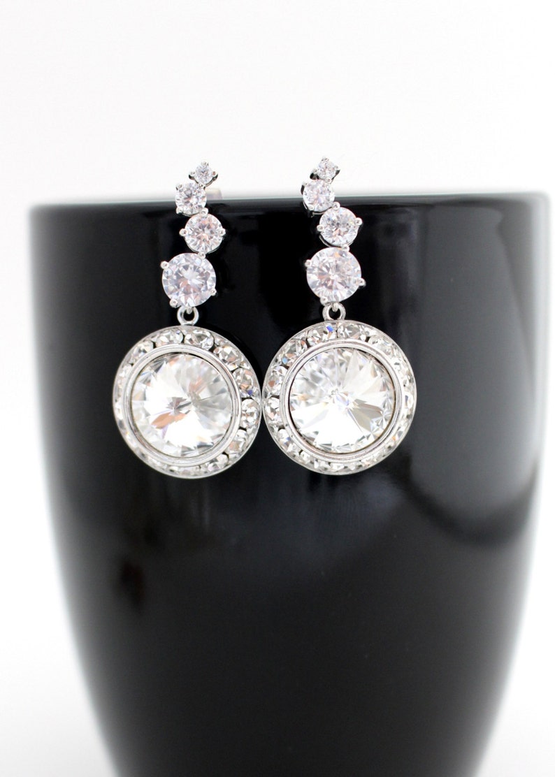 May include: A pair of silver dangle earrings with a large round clear crystal stone at the bottom. The earrings have a row of smaller clear crystal stones above the large stone.