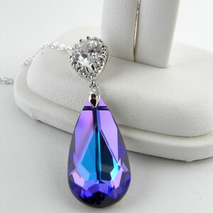 May include: A silver-toned pendant necklace featuring a large, teardrop-shaped, faceted crystal in shades of purple and blue. The crystal is suspended from a smaller, round, clear crystal setting. The necklace is displayed on a white surface.