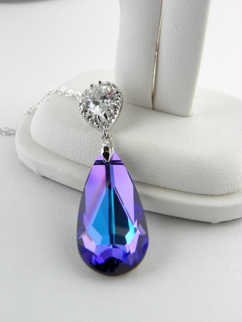 May include: A silver-toned pendant necklace featuring a large, teardrop-shaped, faceted crystal in shades of purple and blue. The crystal is suspended from a smaller, round, clear crystal setting. The necklace is displayed on a white surface.