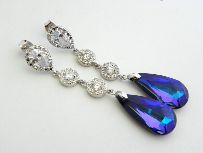 May include: A pair of elegant drop earrings. Each earring features a clear, pear-shaped stone at the top, followed by three round, clear stones. The earrings end with a large, teardrop-shaped, iridescent blue stone.