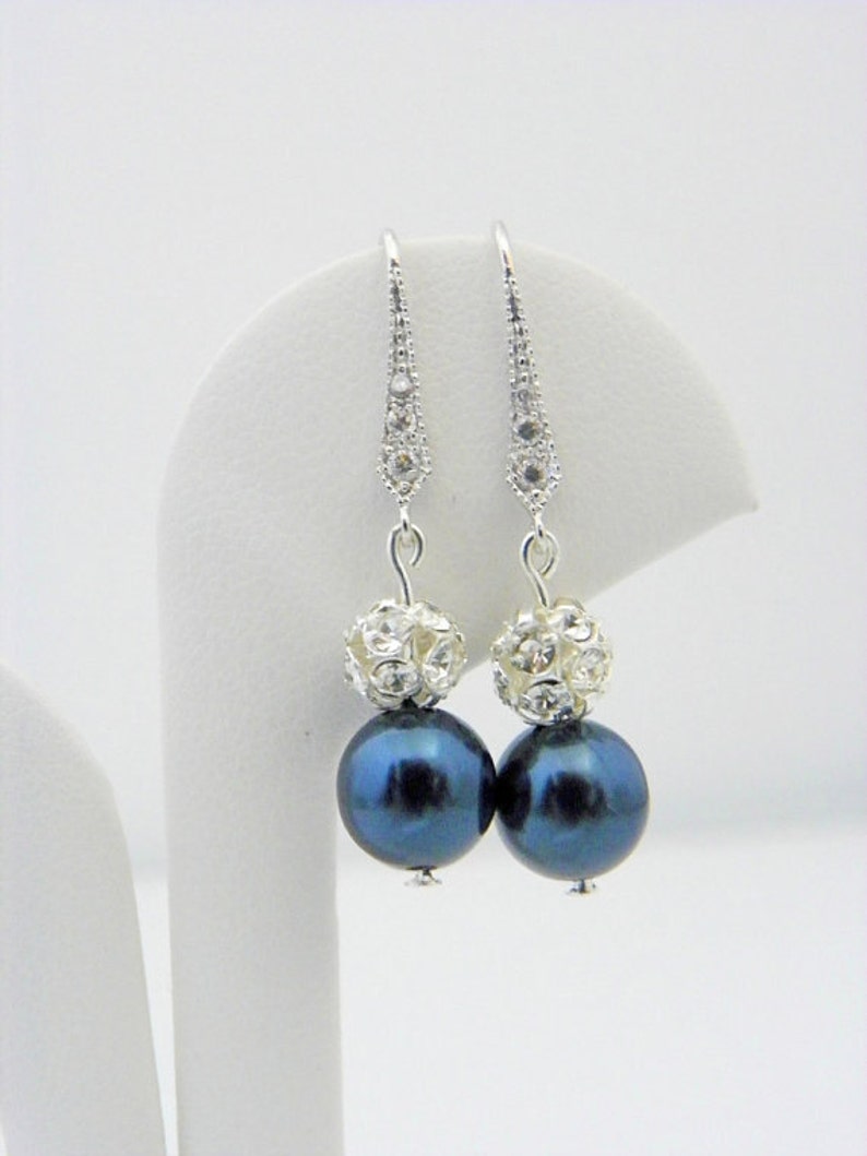 May include: A pair of dangle earrings featuring dark blue, iridescent pearl-like beads. Each earring has a silver-toned, crystal-studded top and a large, round bead. The earrings are displayed against a white background.