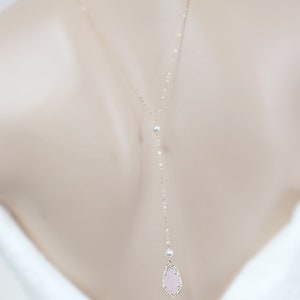 May include: A delicate gold-toned back necklace featuring a pear-shaped, light pink gemstone pendant. The pendant is outlined with small, sparkling crystals. The necklace includes a small pearl accent and a delicate chain that drapes down the back.