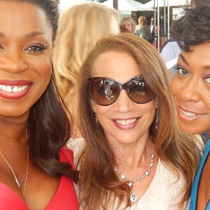 May include: Three women smiling at the camera. One woman is wearing a red dress, another is wearing sunglasses and a white top, and the third is wearing a blue top. They are all outdoors.