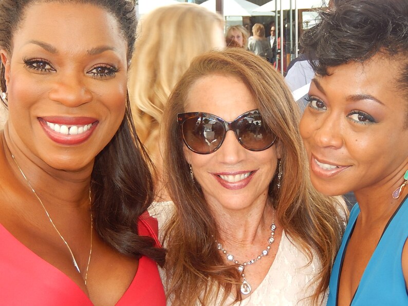 May include: Three women smiling at the camera. One woman is wearing a red dress, another is wearing sunglasses and a white top, and the third is wearing a blue top. They are all outdoors.